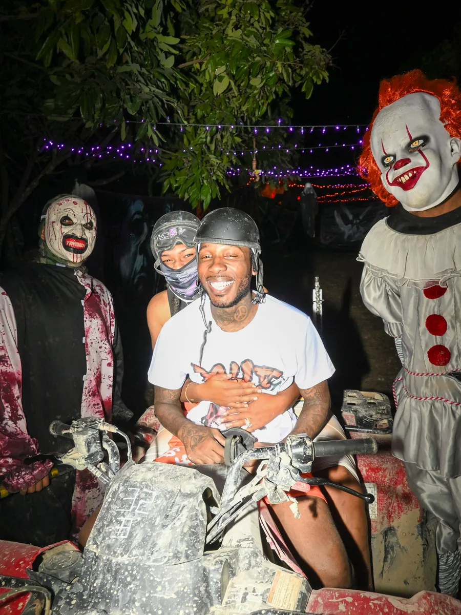 Three riders on an ATV grinning while clowns crowd in around them under string lights