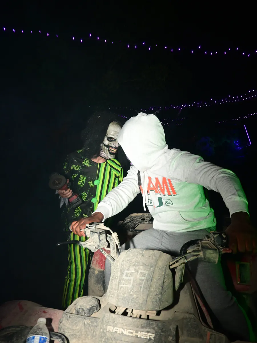 A scare actor in a green striped clown costume leaning in behind a rider sitting on an ATV at night